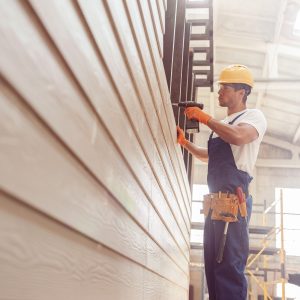 Handsome young man construction worker wearing safety helmet and work overalls while installing exterior wood siding