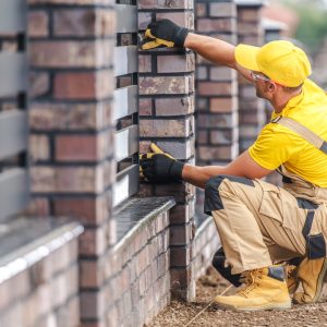 A worker in a yellow shirt and gloves meticulously installs bricks on a stylish fence at a home. The task is performed in bright sunlight, showcasing attention to detail and craftsmanship.