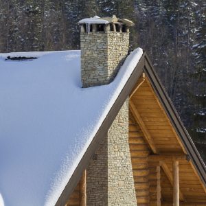 Top of wooden ecological traditional cottage of lumber materials with steep roof, high stone chimney covered with snow on tall spruce tree forest background on bright sunny winter day.