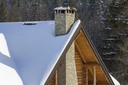 Top of wooden ecological traditional cottage of lumber materials with steep roof, high stone chimney covered with snow on tall spruce tree forest background on bright sunny winter day.